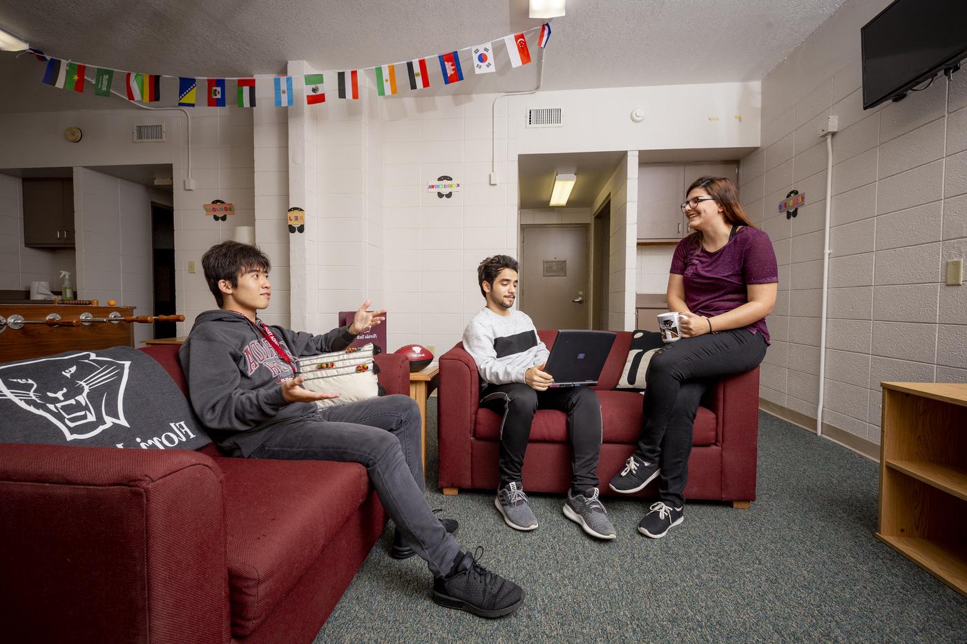 Three students hanging out in a residence hall common area. A TV, foosball table, and couches are visible