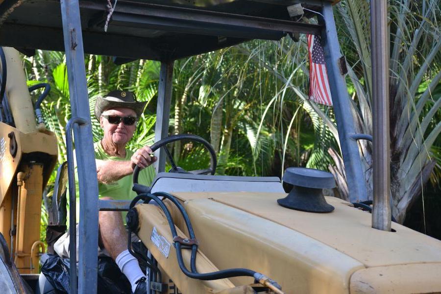 A friendly-looking, mature groundskeeper, Hank Hughes, in a neon green t-shirt, cowboy hat and sunglasses, driving a large tractor.